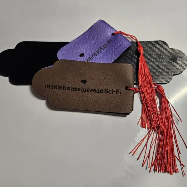Leather bookmarks engraved with 'in this house we read Sci-Fi' in purple, brown, black, and carbon fiber leatherette